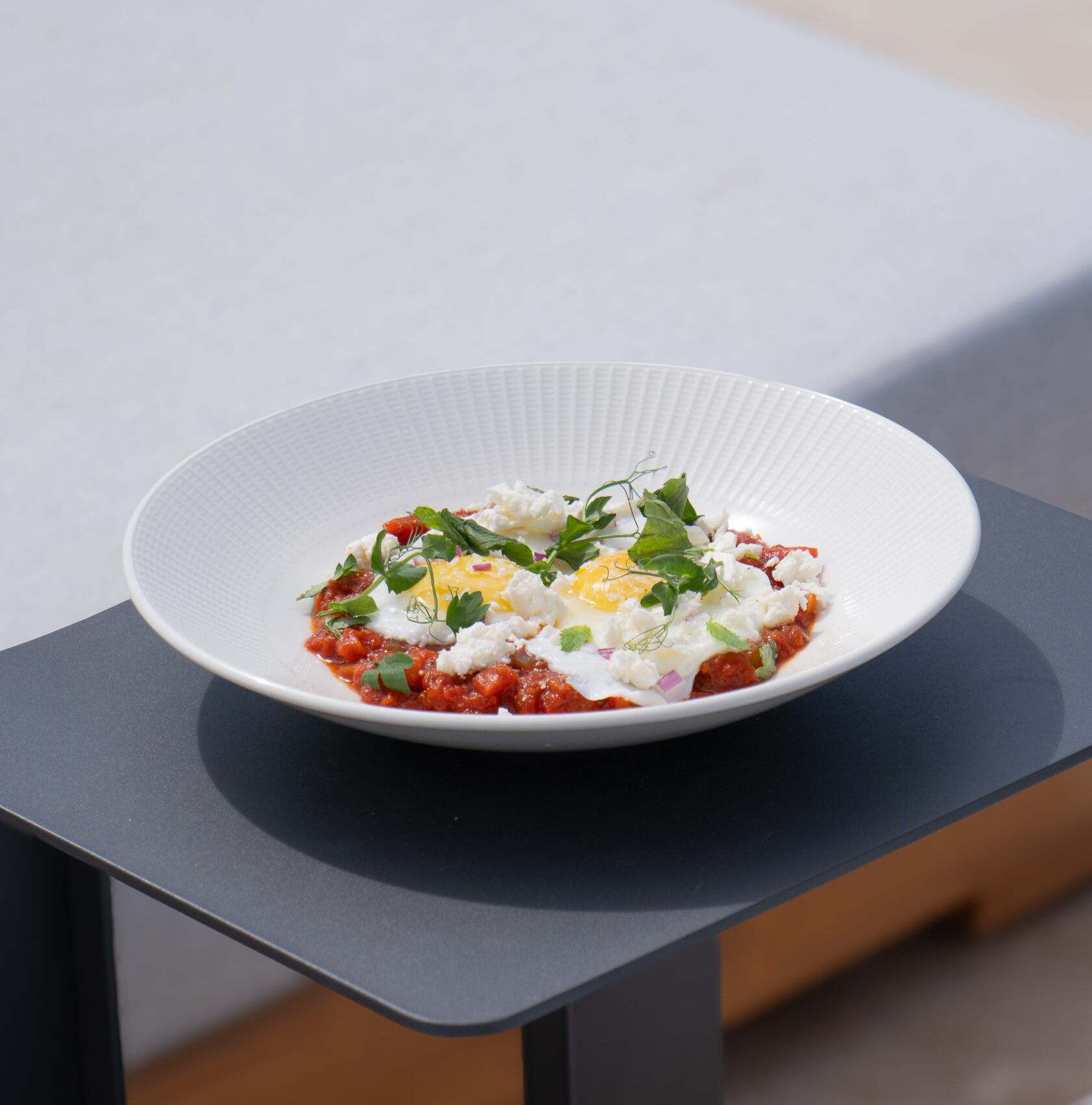 A white plate with shakshuka, featuring poached eggs in a bed of tomato sauce, topped with crumbled cheese and garnished with green herbs, on a black table reminiscent of the elegant dining found at luxury villas in Bermuda.