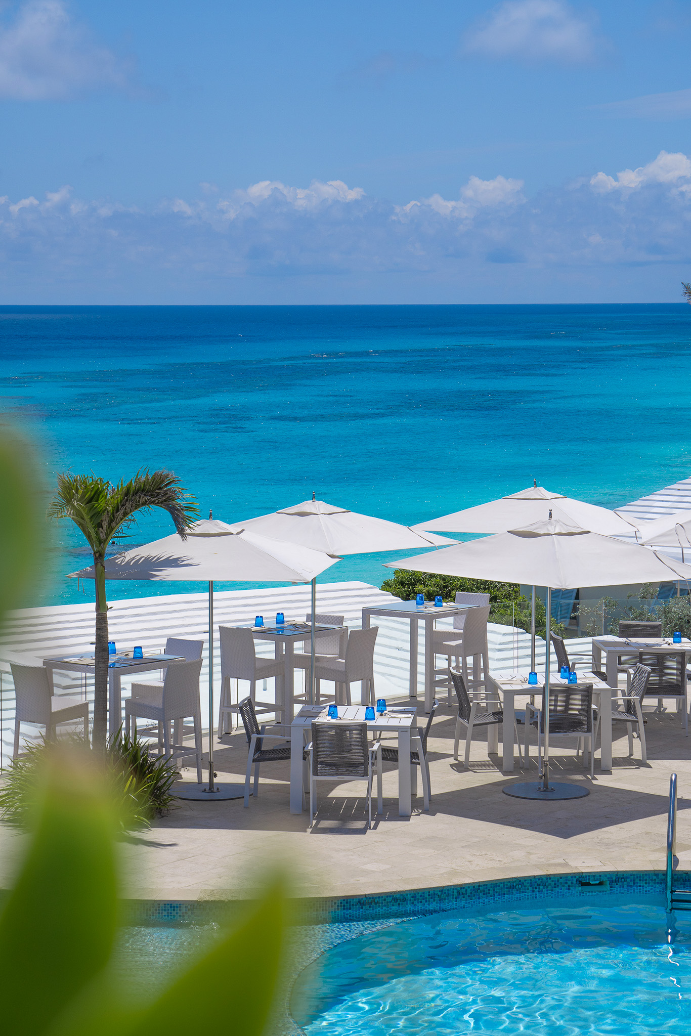 Outdoor dining area with white tables, chairs, and umbrellas overlooking a turquoise ocean and clear blue sky at a luxury hotel in Bermuda.
