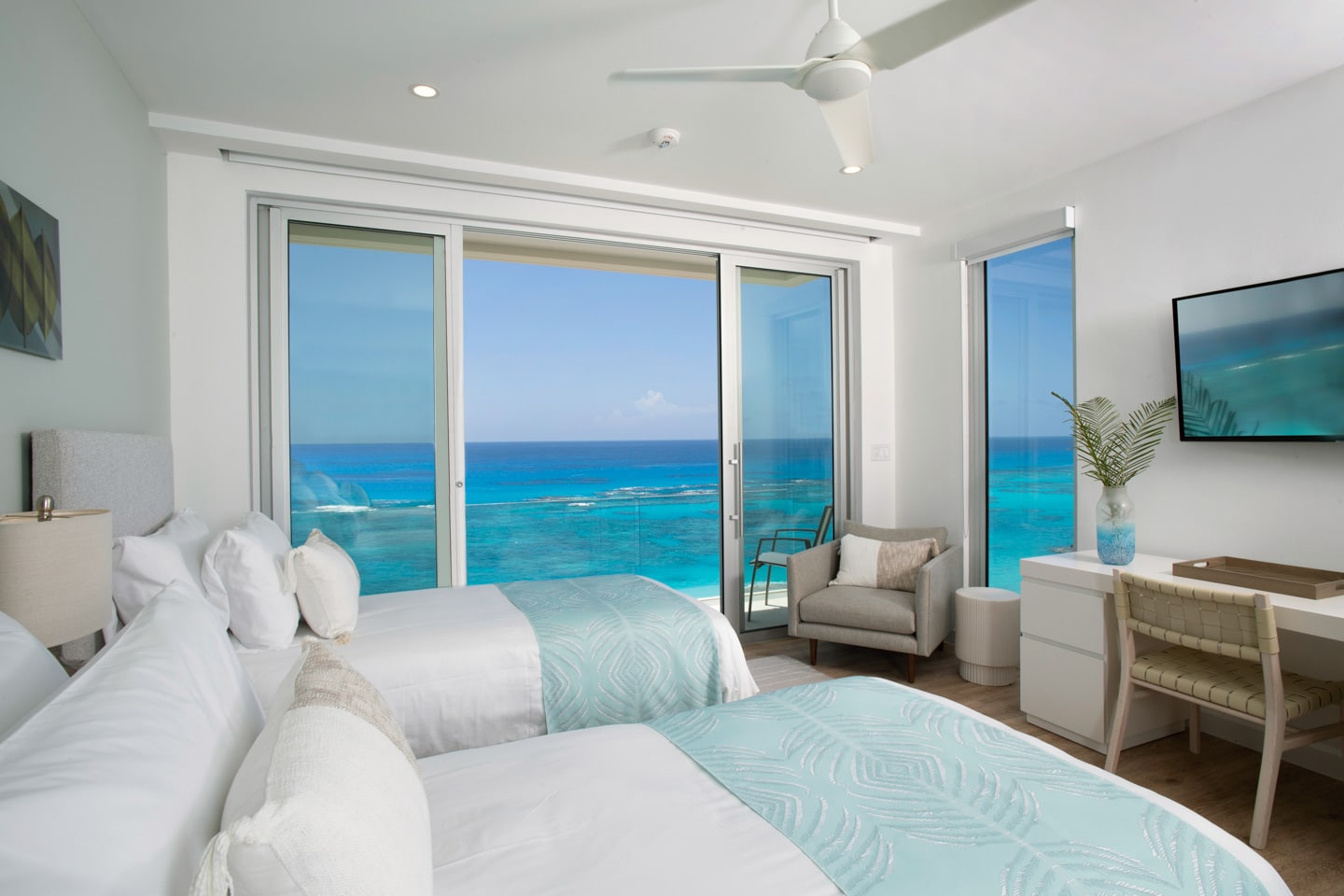A bedroom with two beds, a desk, and glass sliding doors displaying an ocean view.