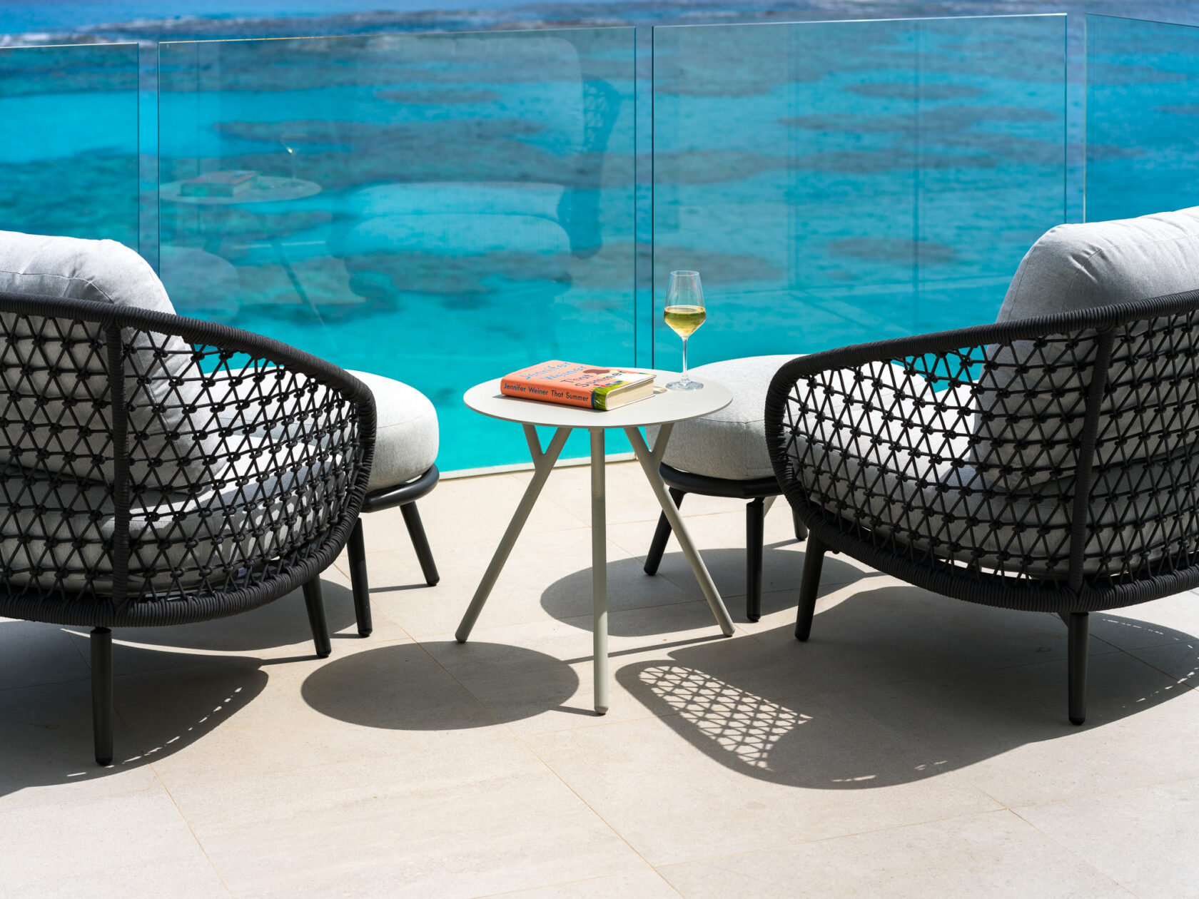 Two wicker chairs face an ocean view at a luxury villa in Bermuda, with a small table between them holding a book and a glass of white wine.