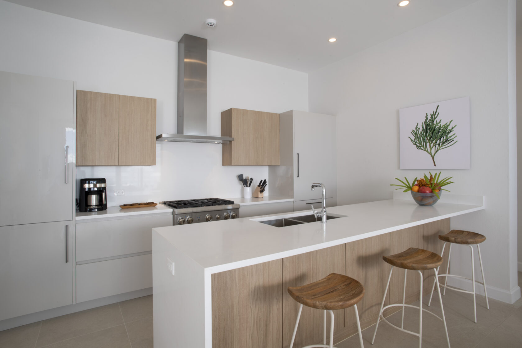 Modern kitchen with white countertops, a stainless-steel stove and hood, light wood cabinetry, three wooden bar stools, and a bowl of fruit on the island. A tree artwork hangs on the wall, elevating this space reminiscent of luxury residences in Bermuda.