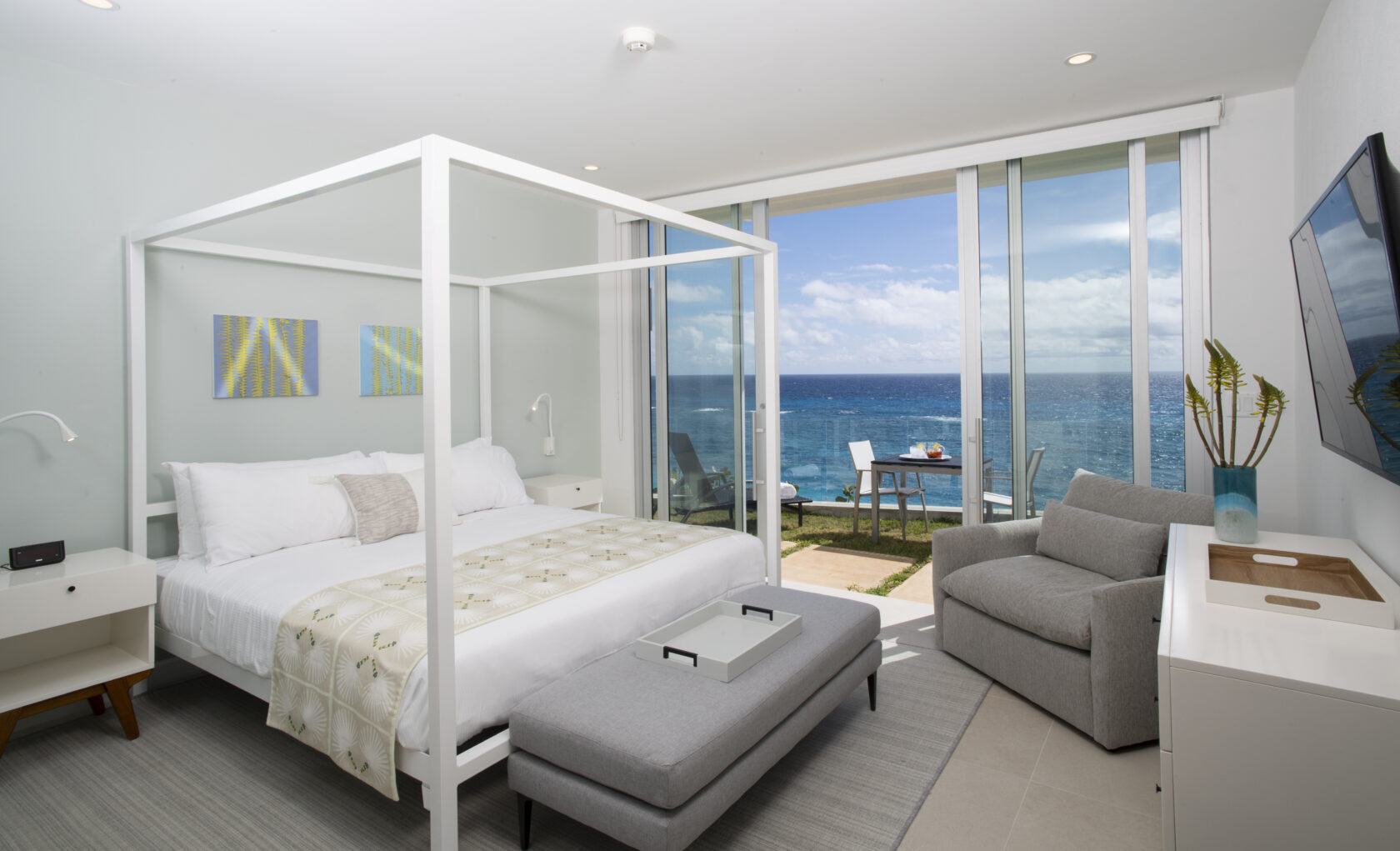 A modern bedroom at this Bermuda boutique hotel features a canopy bed, armchair, and ocean view through sliding glass doors leading to a small balcony. The room is decorated in neutral tones with minimalistic furnishings, capturing the essence of luxury residences in Bermuda.