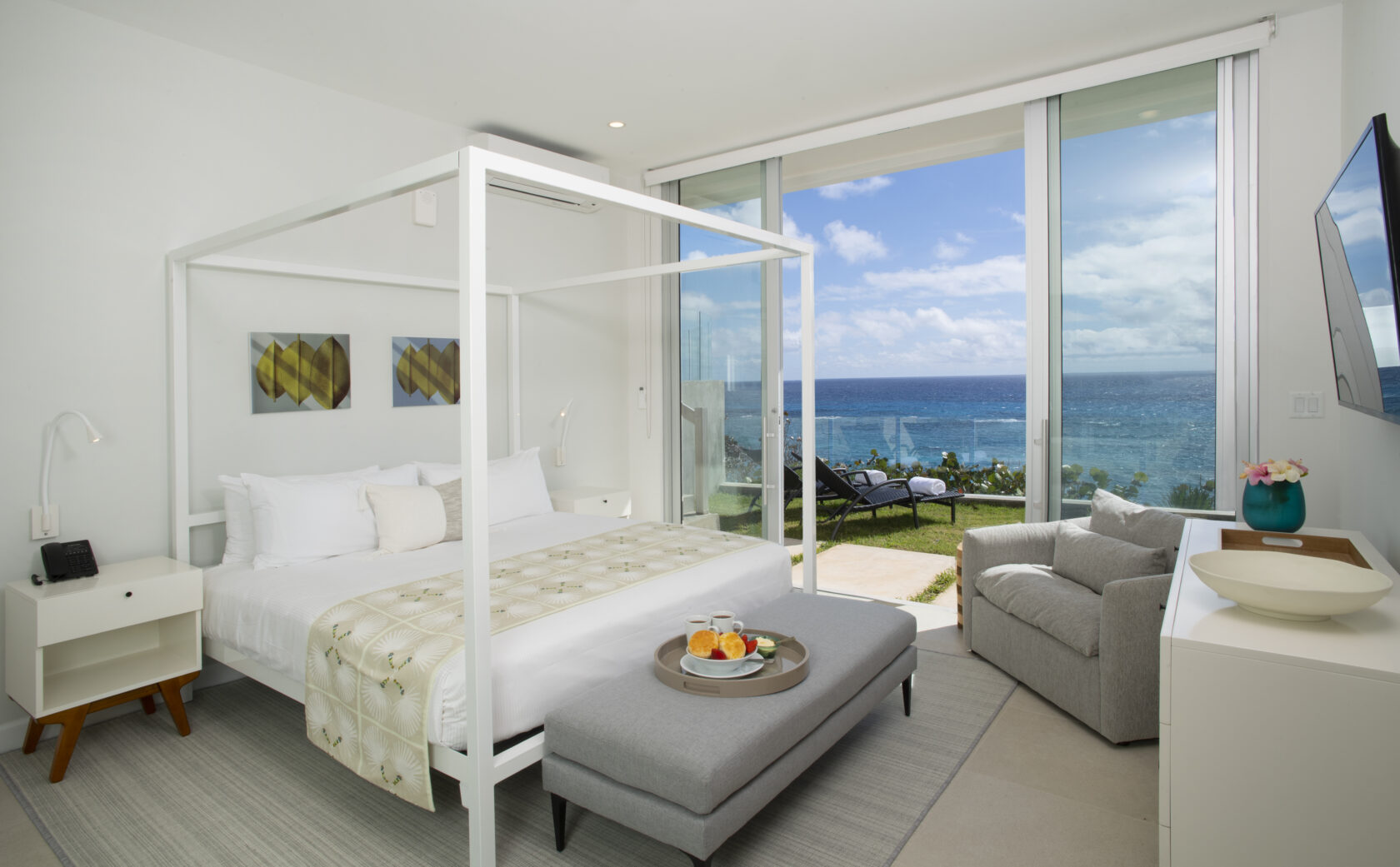 Modern, brightly lit bedroom at a luxury residence in Bermuda, featuring a four-poster bed, ocean view through floor-to-ceiling windows, and minimalist furnishings including a white nightstand, cushioned bench, and armchair.