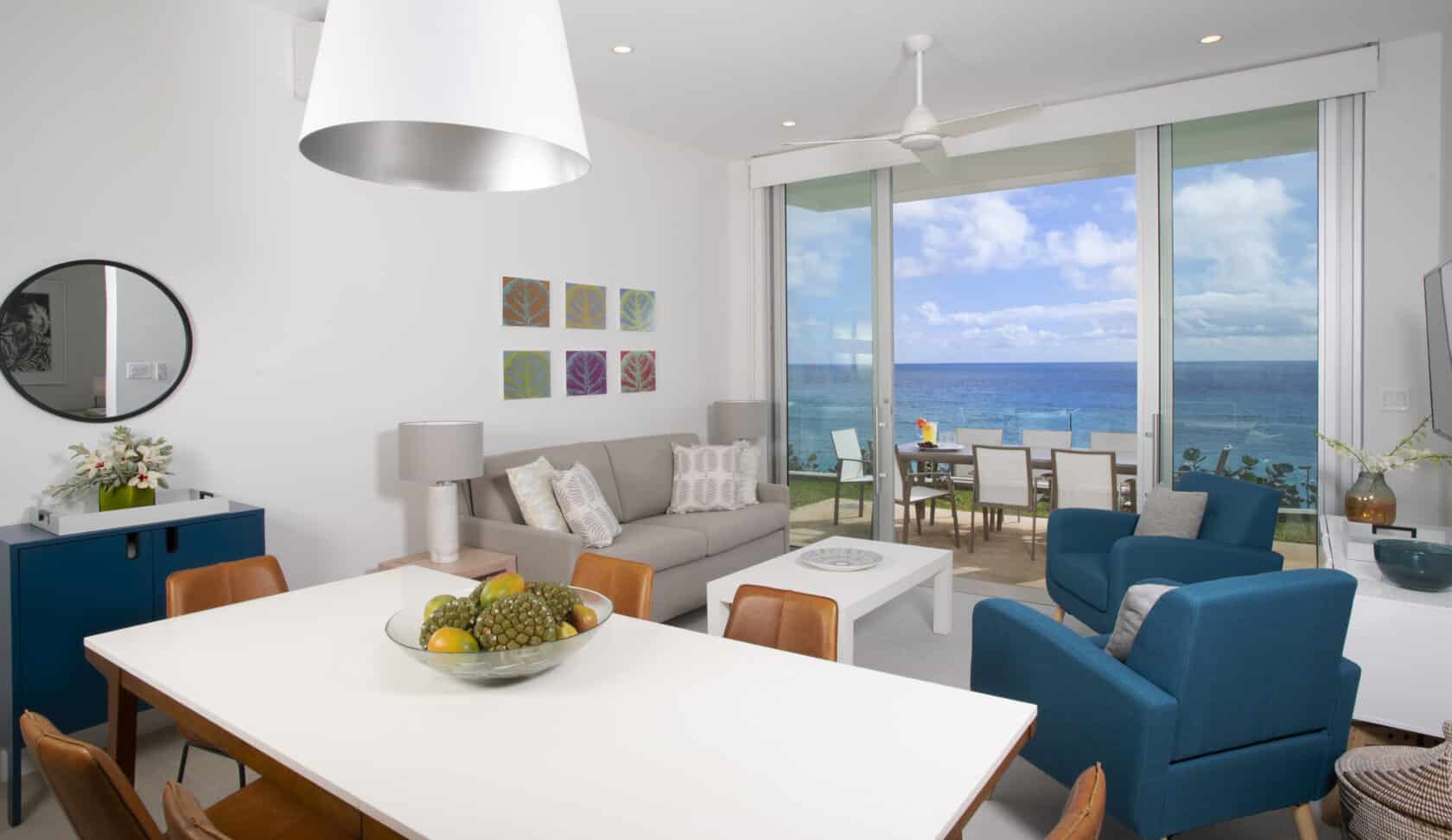 A modern living and dining area in one of the luxury villas Bermuda boasts, featuring a white dining table, blue chairs, a grey sofa, and ocean views through large sliding glass doors leading to a balcony with outdoor seating.