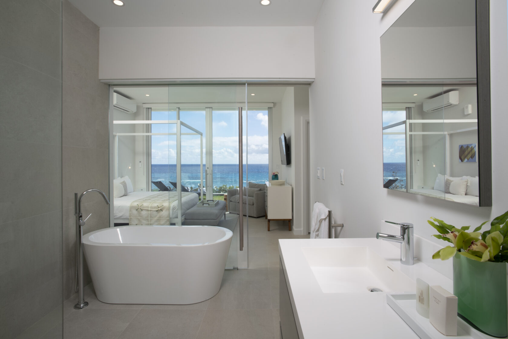 Modern bathroom with a freestanding tub and sink, overlooking a bedroom with floor-to-ceiling glass windows showing an ocean view, typical of luxury villas in Bermuda.