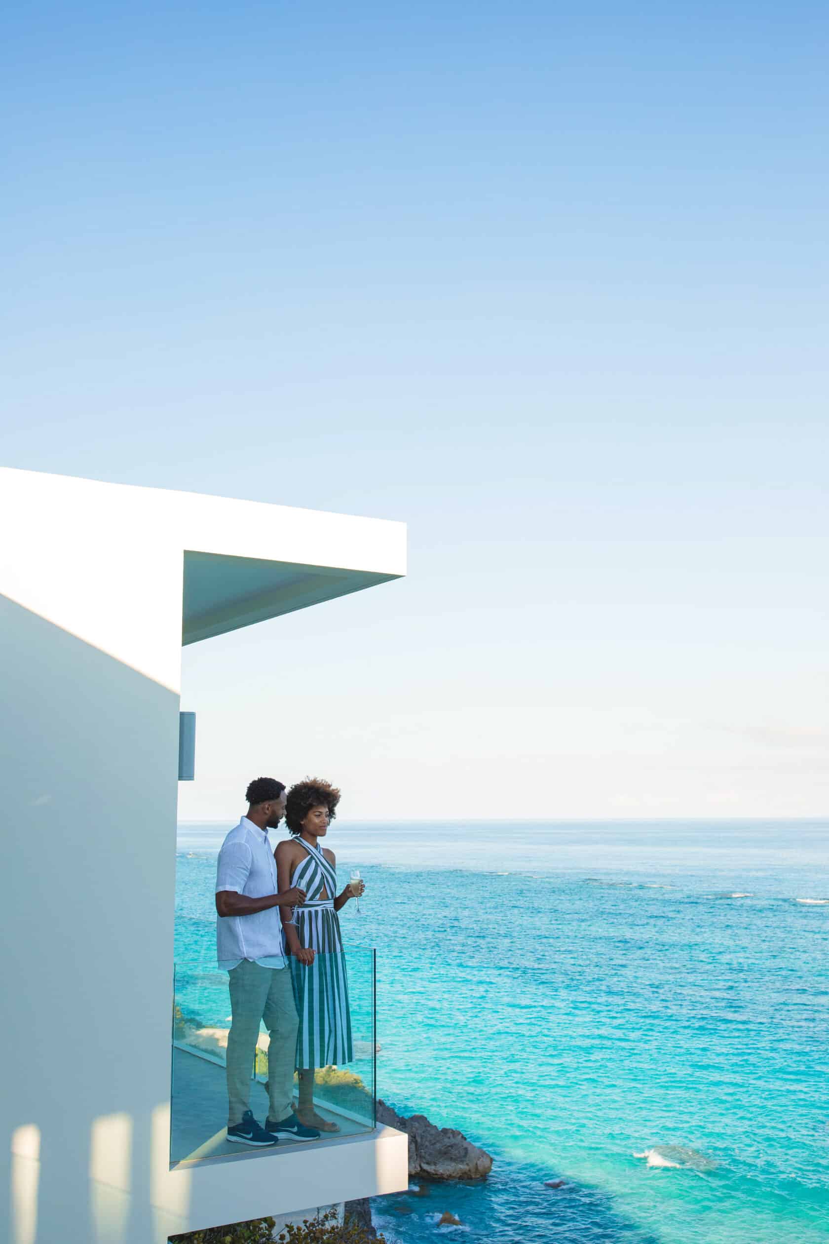 A couple stands on a balcony overlooking a bright blue ocean, with a modern white building visible. The sky is clear and sunny, embodying the serene elegance of luxury residences in Bermuda.