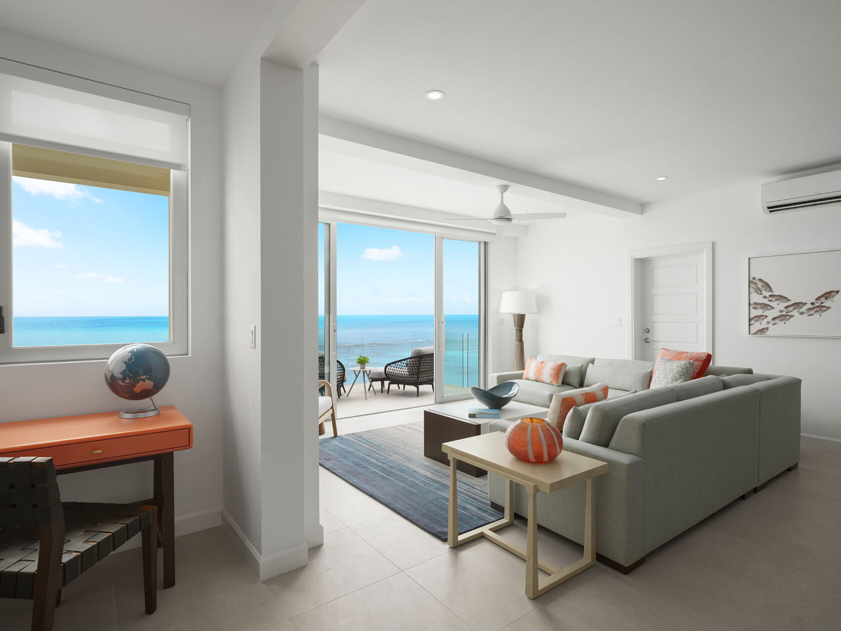 A modern living room with a light gray sectional sofa, a wooden coffee table, and a globe on an orange desk. Large windows and sliding doors offer a clear view of the ocean, reminiscent of luxury villas Bermuda is renowned for.