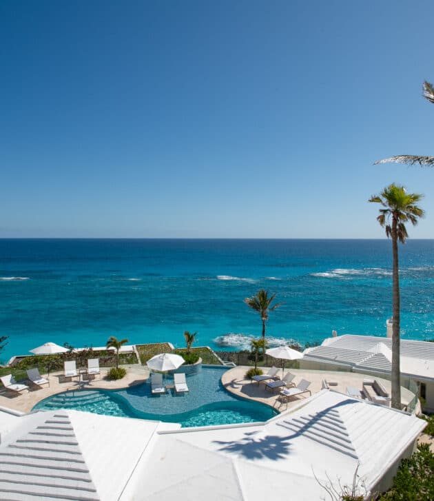 An aerial view of an outdoor pool with a poolside seating area, all overlooking an ocean.