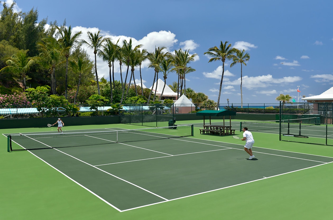 Two people playing tennis at a tennis court with palm trees in the background.