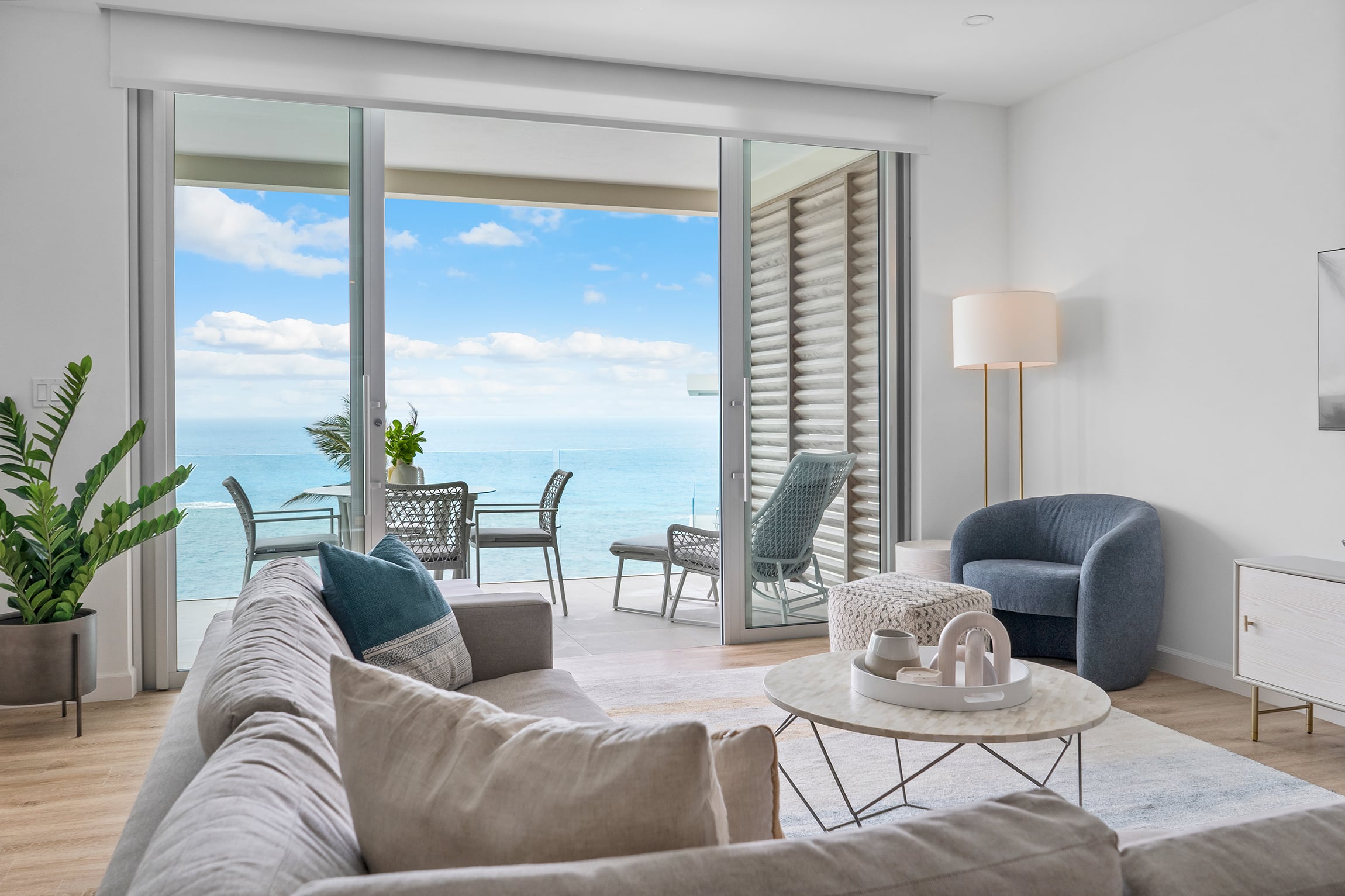 A modern living room with a gray sofa, round coffee table, blue armchair, and glass doors opening to a balcony overlooking the ocean. The scene evokes the sophistication found in luxury villas Bermuda, complete with outdoor furniture and a potted plant on the balcony.