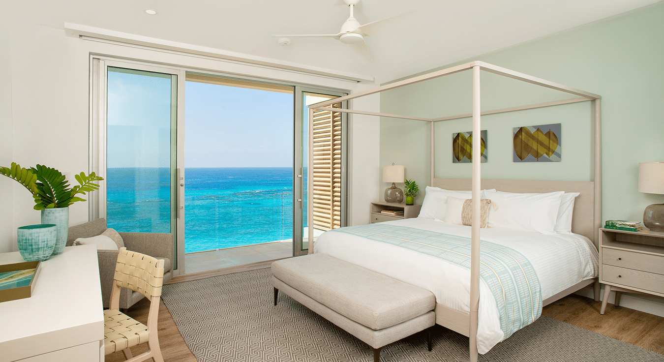 A modern bedroom with a canopy bed, desk, and balcony overlooking a clear blue ocean in a Bermuda boutique hotel. The room has light green walls and a ceiling fan.