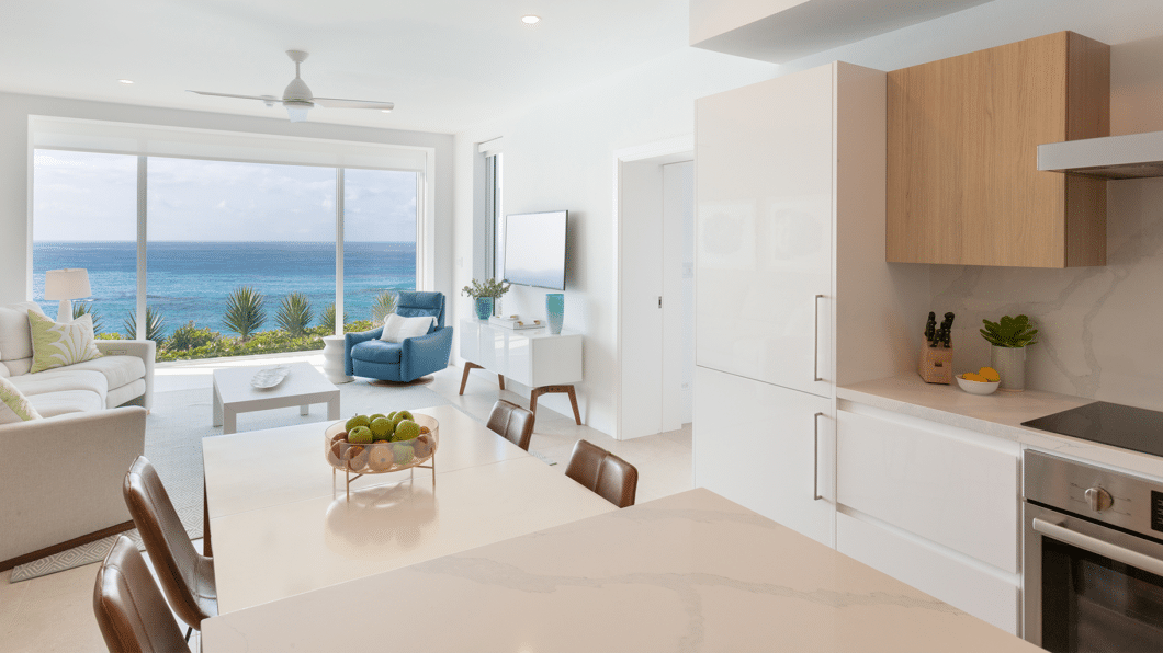 Modern kitchen and living room with ocean view in a Bermuda boutique hotel. White and wood finishes, stainless steel appliances, dining table with chairs, and lounge area with light-colored sofas and a blue armchair.