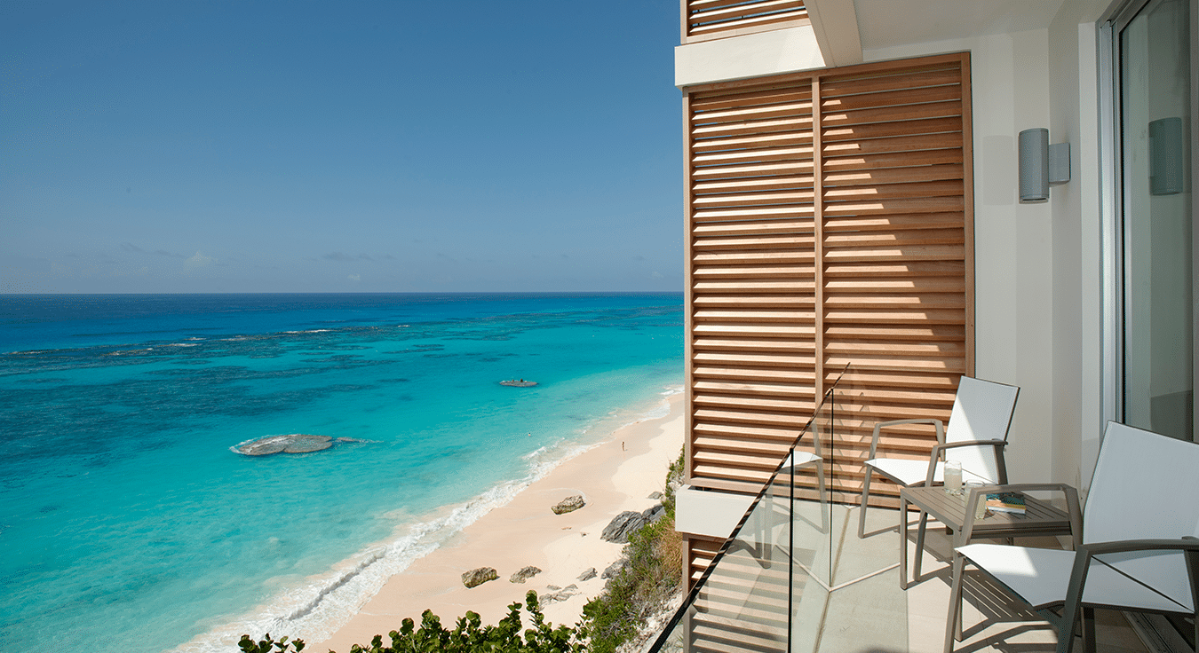 A balcony with two chairs overlooks a beach with turquoise water, white sand, and scattered rocks under a clear blue sky, embodying the serene beauty found at luxury residences Bermuda.