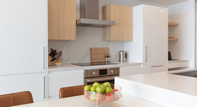 Modern kitchen with white cabinets, wood accents, a stovetop, oven, and a countertop with a bowl of green and red apples. Kitchen appliances include a kettle and a cutting board. Inspired by the sleek designs of luxury villas in Bermuda, this space exudes both elegance and functionality.
