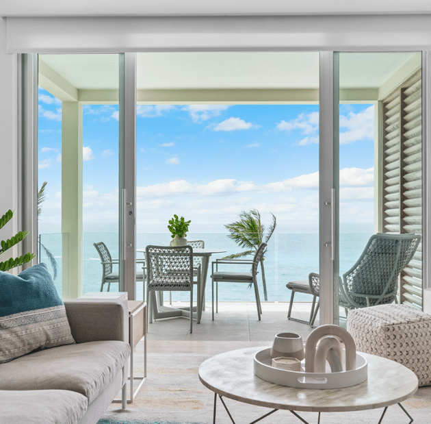 Modern living room with a view of a spacious balcony featuring outdoor furniture and potted plants, overlooking a calm ocean under a partly cloudy sky, reminiscent of the serene atmosphere found in luxury residences Bermuda.