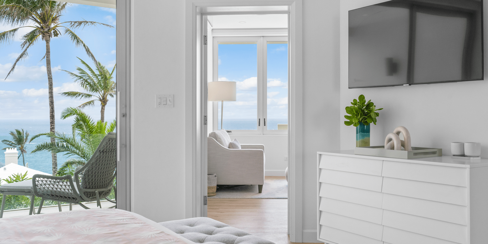 A bright bedroom with a view of the ocean through an open doorway to a balcony. The room, part of one of the luxury residences in Bermuda, has a TV on the wall, a white dresser with a plant, and a seating area in front of wide windows.