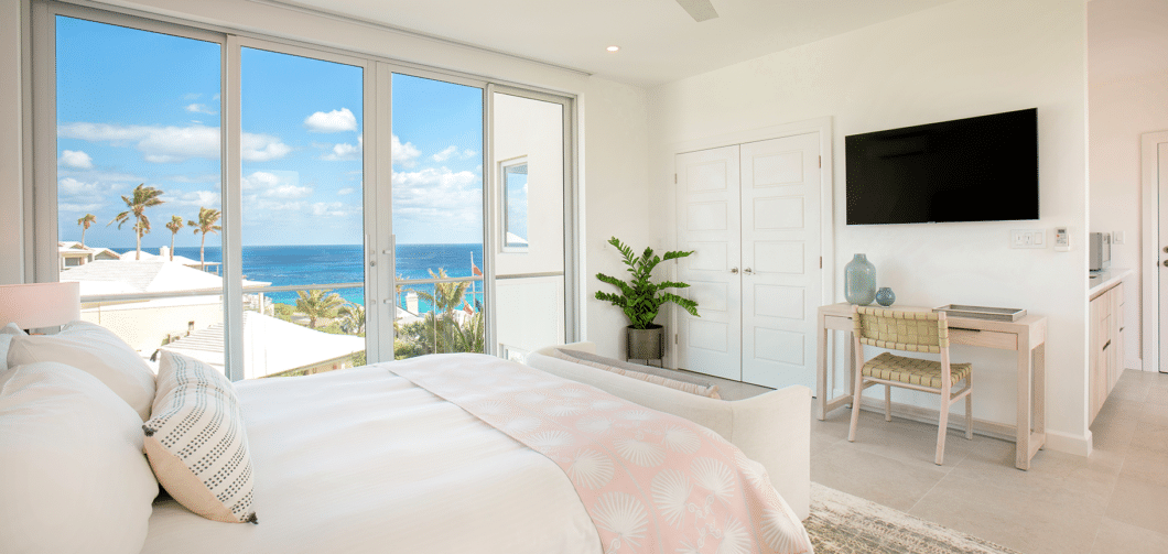 A modern bedroom in this luxury hotel Bermuda features a large bed, a desk with a chair, potted plants, a wall-mounted TV, and large windows with an ocean view.