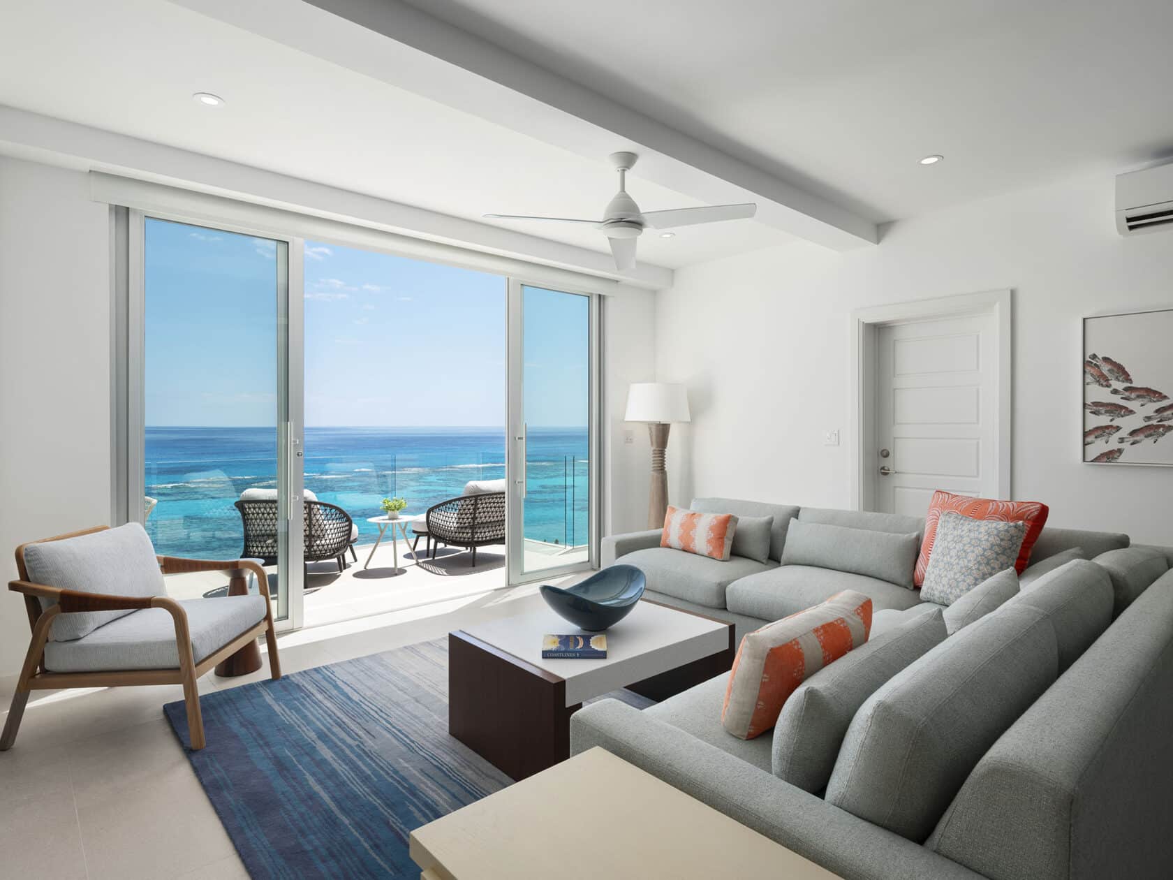 Modern living room with light gray furniture, a coffee table, and a blue rug. A large sliding glass door opens to a balcony with chairs, overlooking the ocean. A ceiling fan is centered above. Ideal for those seeking luxury residences in Bermuda, this space epitomizes elegance and comfort.