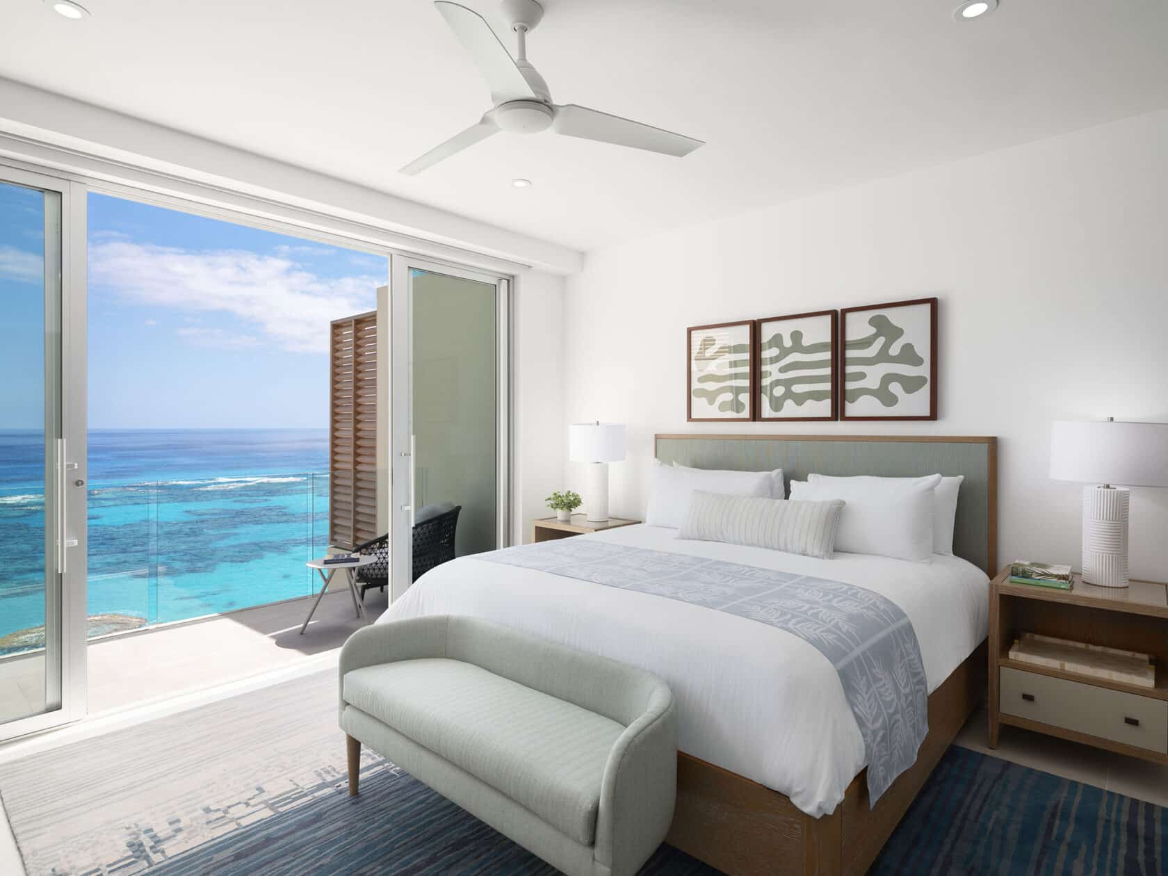 Modern bedroom with a large bed, white bedding, a mint green bench, and ocean view through sliding glass doors. A ceiling fan and wall art add decor, and natural light fills the space. Experience it all at one of the luxury villas Bermuda is renowned for.