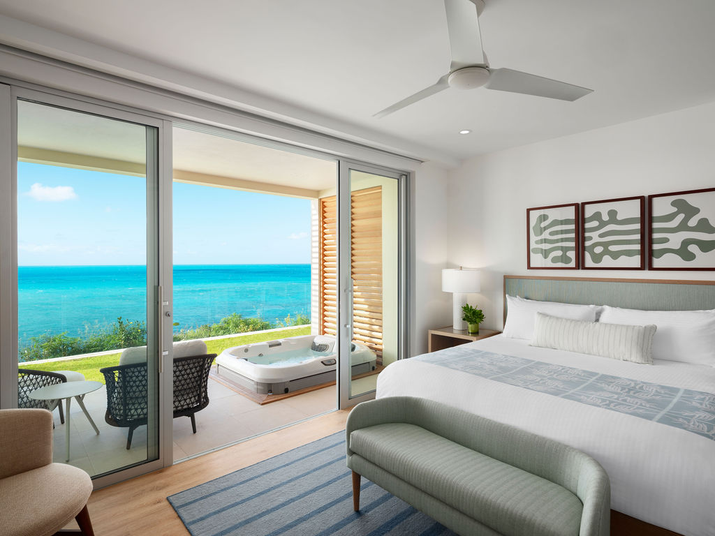 Modern bedroom in a luxury hotel in Bermuda with a large bed, sliding glass doors opening to a balcony with ocean views, outdoor seating, and a hot tub. Room features a ceiling fan, framed wall art, and light furnishings.