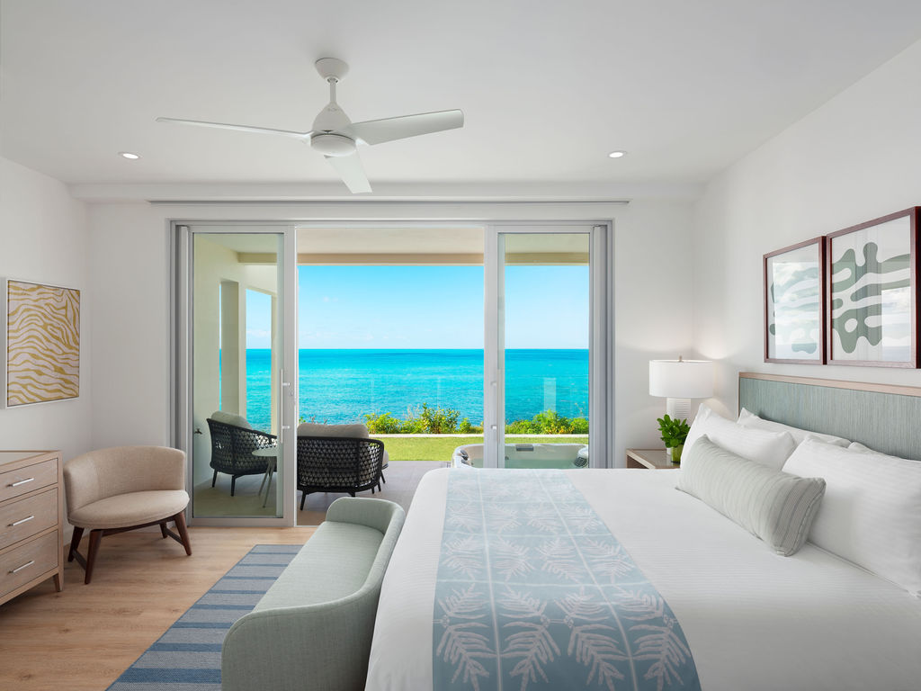 A modern bedroom with a large bed, armchair, and dresser. The room features a ceiling fan, two framed artworks, and a sliding glass door opening to a balcony with an ocean view, reminiscent of luxury residences in Bermuda.