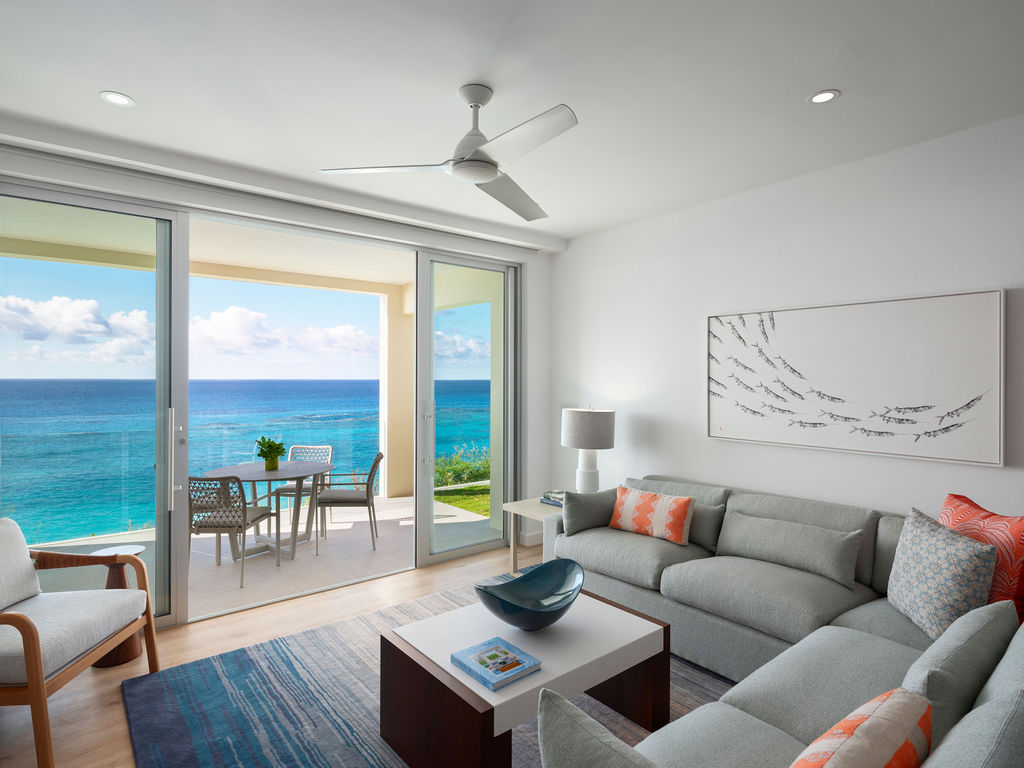 A modern living room with gray furniture and orange accents, reminiscent of luxury residences in Bermuda, adjacent to a balcony overlooking the ocean. The room features a ceiling fan, a coffee table, and large sliding glass doors.
