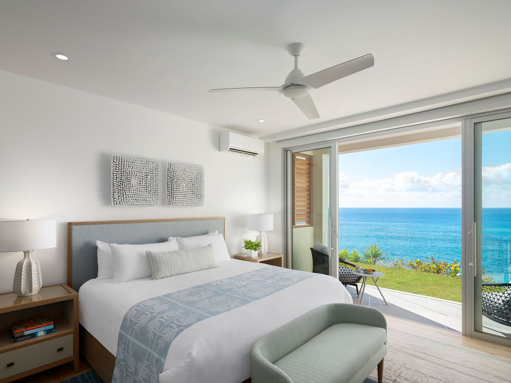 Modern bedroom with a large bed, armchair, bedside tables, and table lamps. Floor-to-ceiling windows open to a balcony with ocean views. Room features a ceiling fan and minimalist decor, reminiscent of luxury residences in Bermuda.