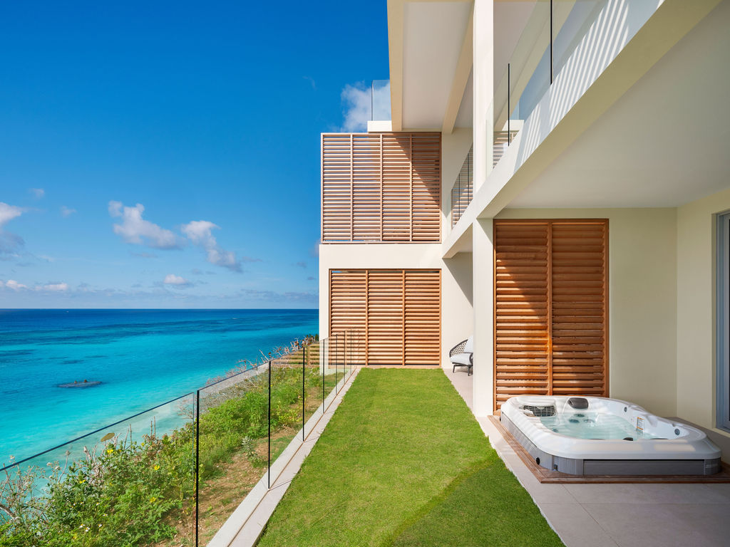 A modern beachfront building with a glass railing, green lawn, and a hot tub on the patio overlooking a clear blue ocean under a bright, sunny sky, creating the perfect setting for one of the finest bermuda boutique hotels.