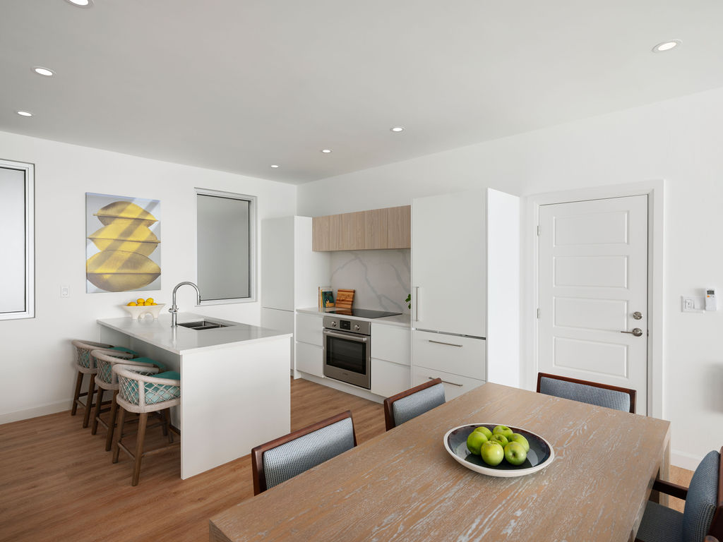 Modern kitchen and dining area featuring white cabinets, stainless steel appliances, a wooden dining table with a fruit bowl, and a kitchen island with barstools. Contemporary art is hung on the wall, reminiscent of the elegance found in luxury residences Bermuda boasts.